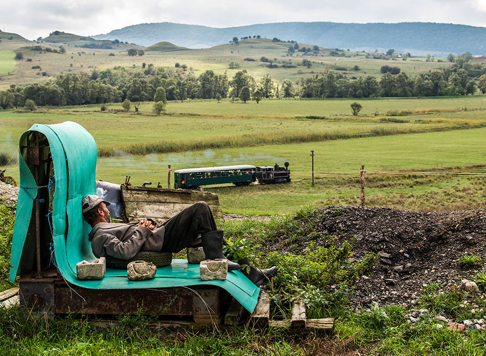 Mann liegt auf einer selbstgebauten Bank und schaut auf einen im Tal vorbeifahrenden Zug